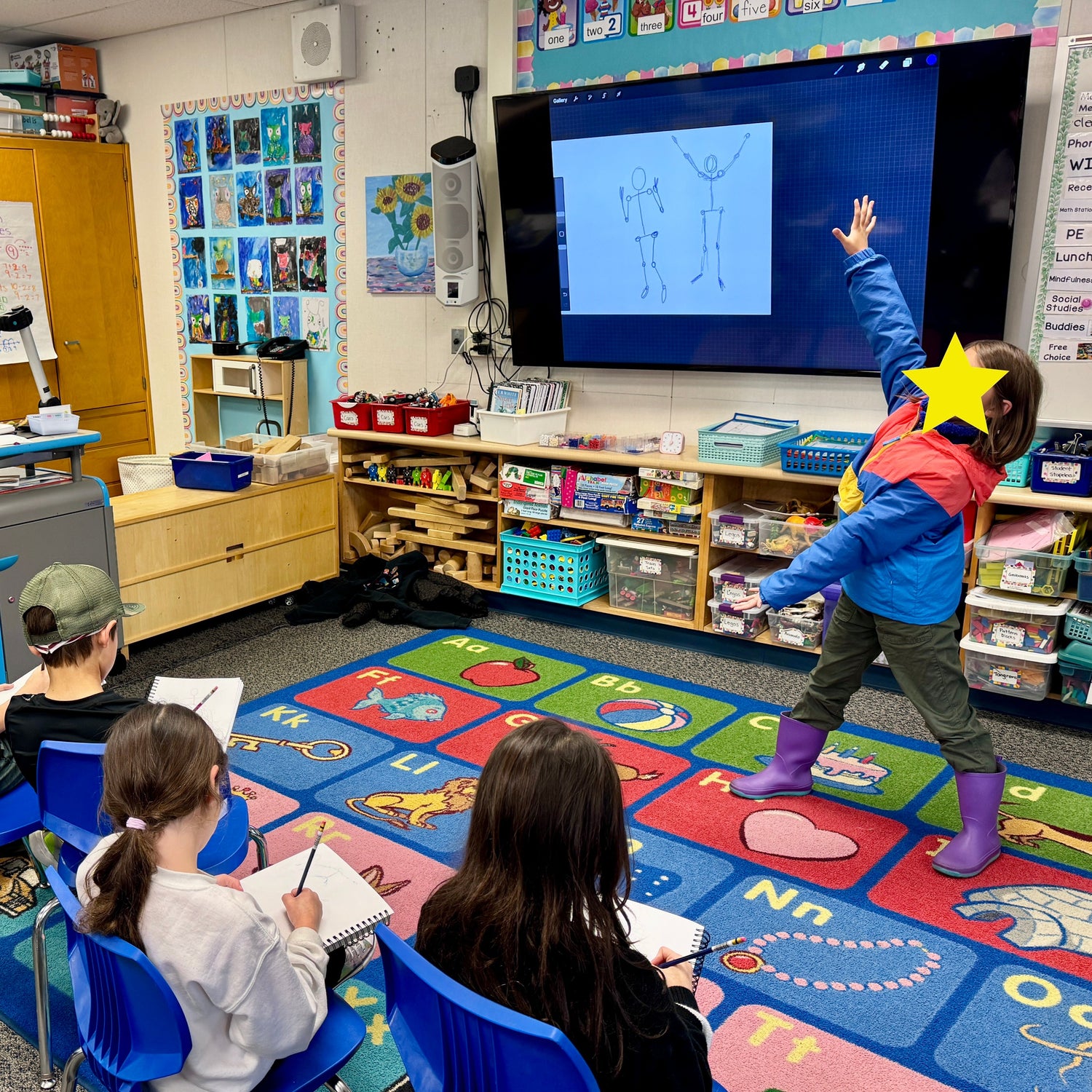 Classroom with children sitting on chairs, facing a large screen displaying educational content.